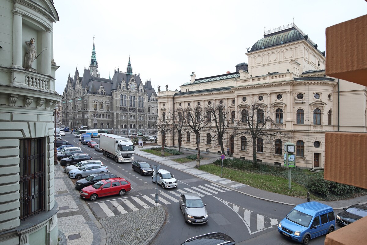 The image captures a street view featuring a bustling roadway lined with parked cars. On one side, the elegant architecture of the historic Liberec City Hall and Šaldovo Theatre can be seen, showcasing ornate detailing and spires, contributing to the city's cultural atmosphere.