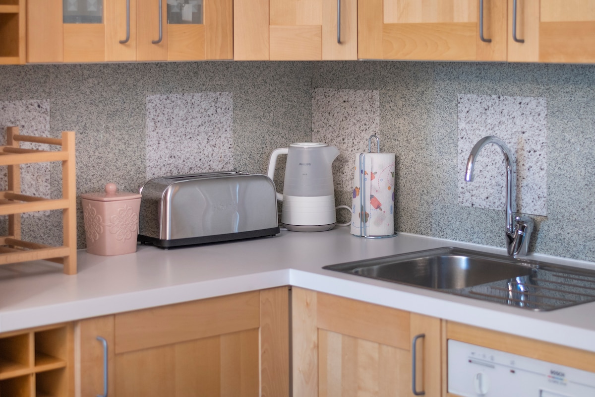 The kitchen area features light wooden cabinetry and a white countertop. A kettle, toaster, and decorative container are neatly arranged. A stainless steel sink and a modern faucet are visible, complemented by the textured grey backsplash that adds subtle contrast.