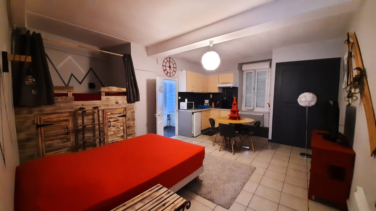 A warm studio space is seen, featuring a red bed and a dining area with a round table and black chairs. The kitchen is equipped with light cabinetry and modern appliances. Bright light filters through multiple windows, with neutral tiles underfoot.