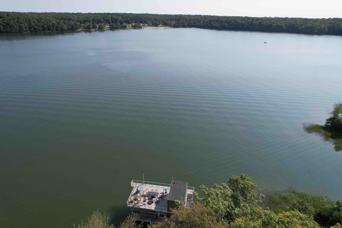 A wide view of the peaceful lake is presented, showcasing calm waters that reflect the surrounding greenery. In the foreground, a private dock extends into the water, featuring a lounging area and clear access to the lake for swimming and recreational activities.