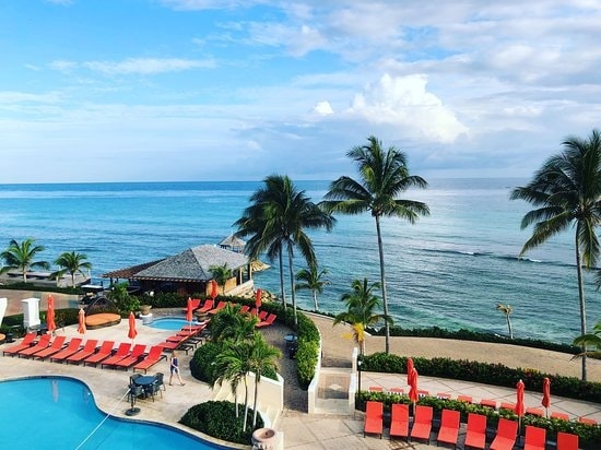 A stunning view of the ocean is captured, showcasing clear blue waters meeting the sky. Palm trees line the shore and a pool area featuring red lounge chairs offers a relaxing space. A thatched-roof cabana is visible near the water's edge.