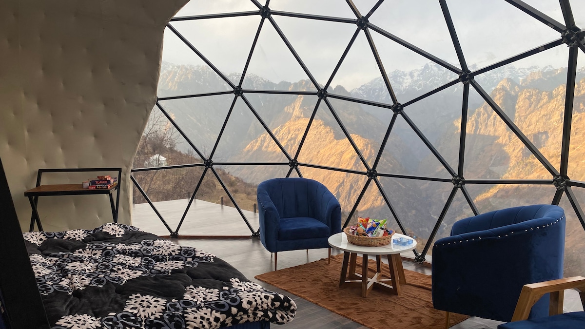 The interior of the dome provides a spacious area with large, geodesic windows offering stunning views of the surrounding mountains. Two blue chairs and a small coffee table are positioned for relaxation, while a comfortable bed adorned with a patterned blanket invites rest.