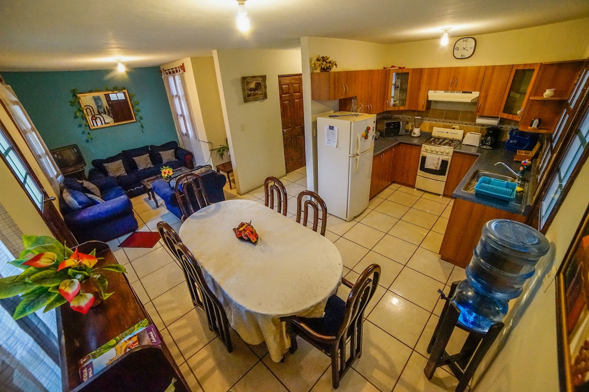 An open kitchen and dining area is visible, featuring a table surrounded by six chairs. A refrigerator and stove are located in the kitchen, with wooden cabinets above and ample counter space. A cozy seating arrangement with a blue sofa is positioned to the side, creating a welcoming atmosphere.