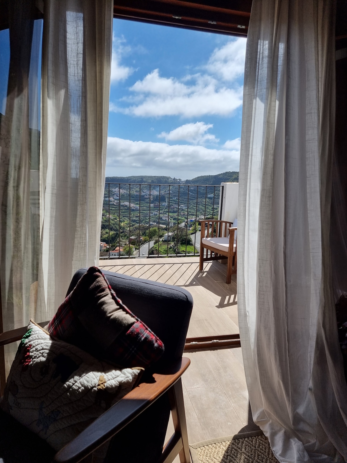A view from the interior space showcasing a seating area with a chair and decorative pillows. Sheer curtains frame the entryway, allowing natural light and a glimpse of the outdoor terrace and surrounding landscape under a partly cloudy sky.