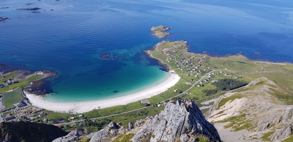 An aerial view captures a stretch of sandy beach bordered by clear turquoise waters. The shoreline is lined with green hills and residential areas, visible in the distance. The calm sea reflects the sky, enhancing the serene atmosphere of this coastal environment.