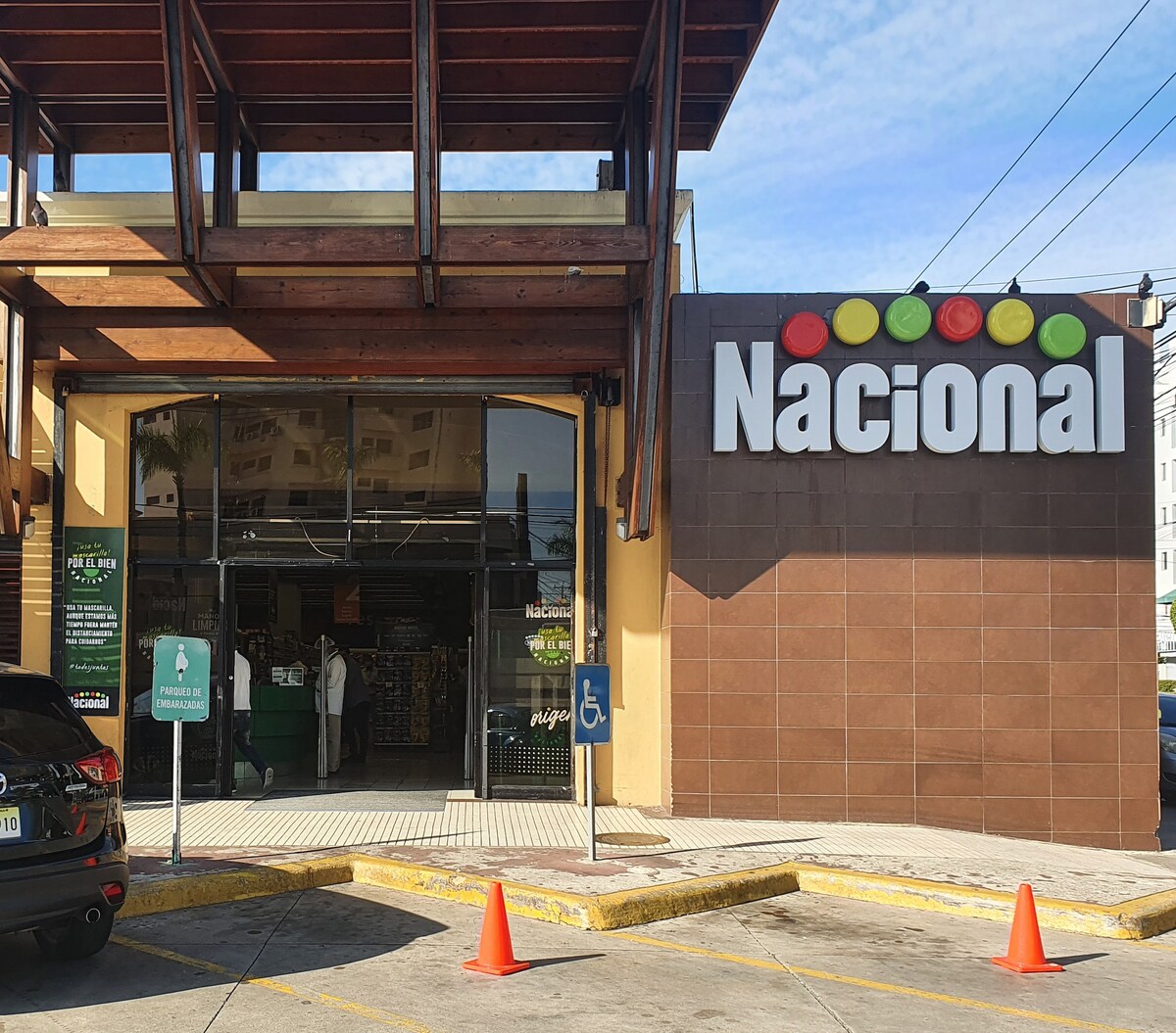 The storefront of a Nacional supermarket is featured, showcasing a modern design with wooden overhangs and large glass doors. Bright signage displays the name 'Nacional' prominently. Accessibility features are visible with a parking space labeled for handicapped access. Orange cones mark the parking area.