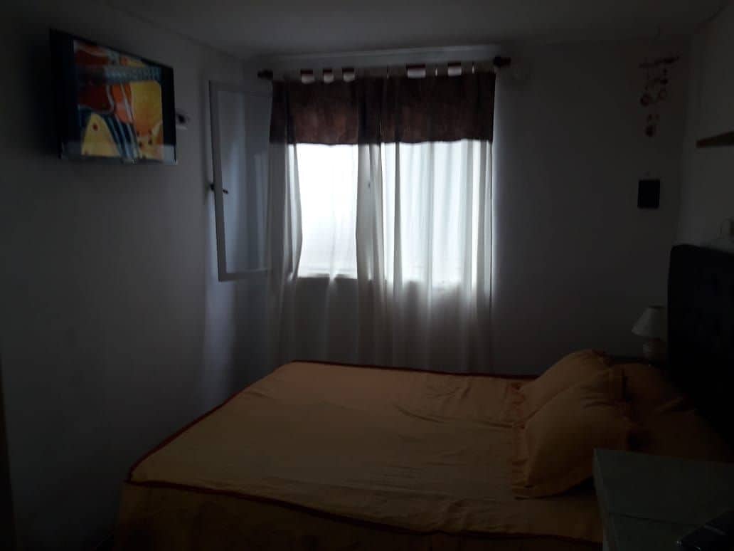 A cozy sleeping area features a spacious bed dressed in a yellow coverlet, complemented by matching pillows. A window draped with sheer curtains allows light to filter in, and a small television is mounted on the wall opposite the bed.