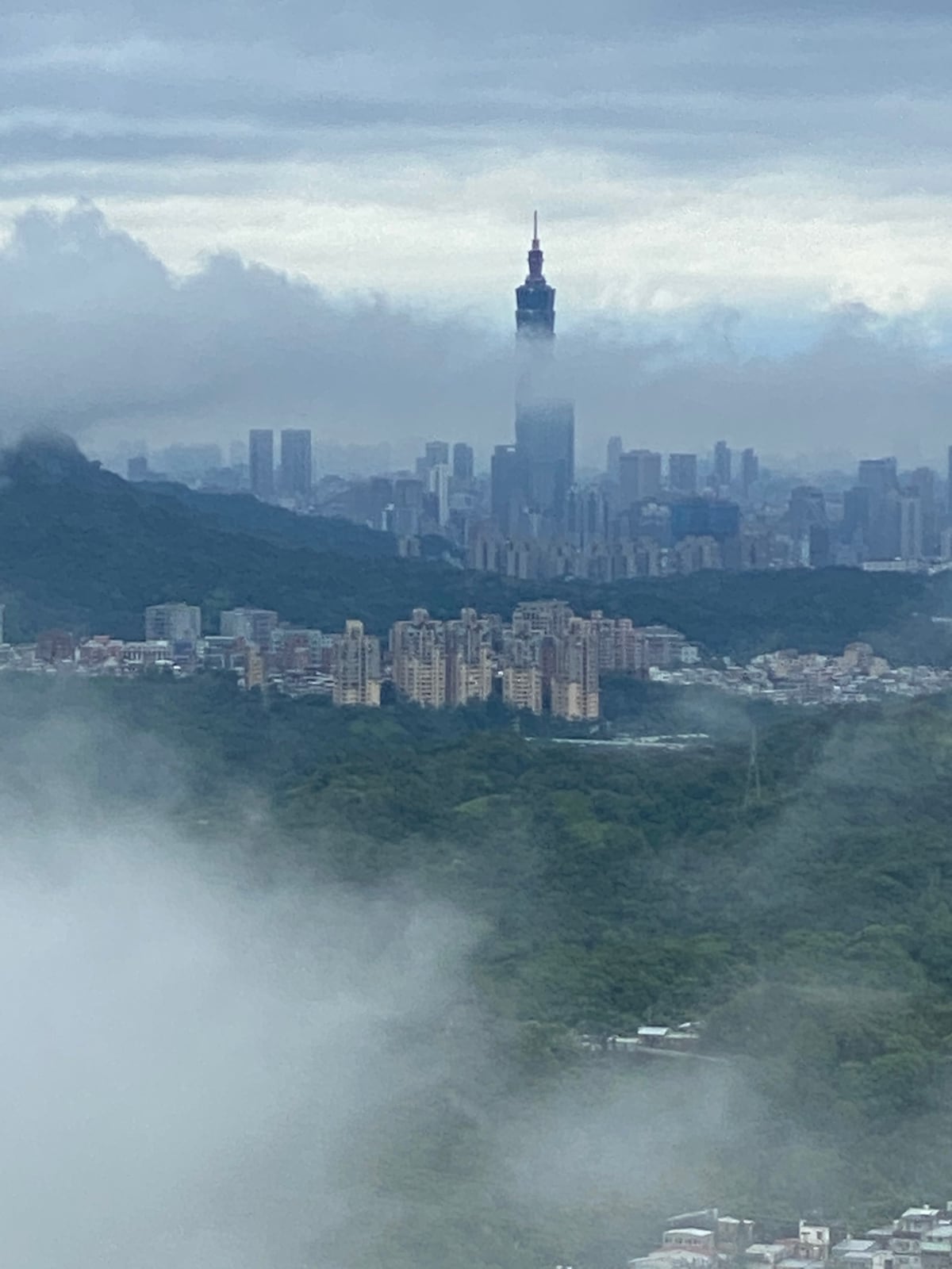 A panoramic view showcases the Taipei skyline, featuring the iconic Taipei 101 partially shrouded in mist. Lush green hills rise in the foreground, contrasting with the urban landscape of high-rise buildings that extend into the distance, framed by soft clouds.