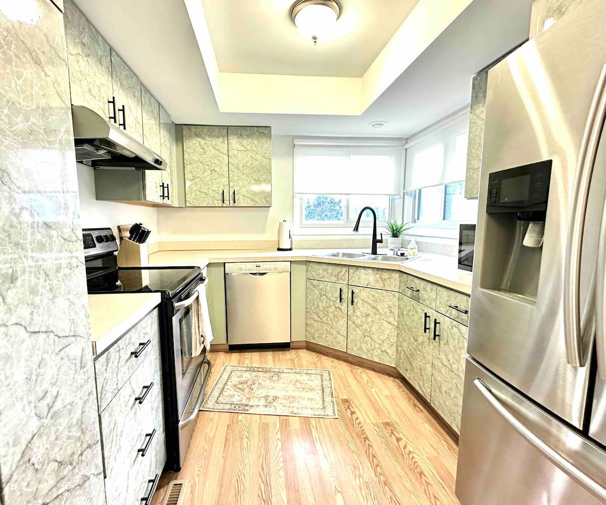 A well-equipped kitchen features modern appliances, including a stainless steel refrigerator and stovetop. Light-colored cabinetry with a marble finish complements the hardwood flooring. Large windows allow natural light to illuminate the space, while a subtly patterned rug adds warmth underfoot.