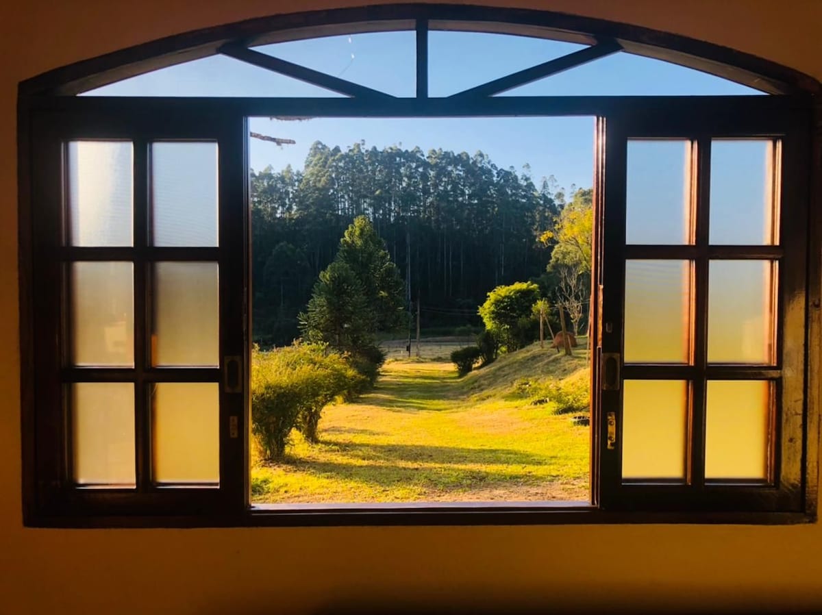 An arched window features multiple wooden panes, opening to a serene landscape. A pathway lined with green grass and bushes extends towards a tree-lined backdrop. Natural light brightens the interior space, enhancing the view of the vibrant outdoors.