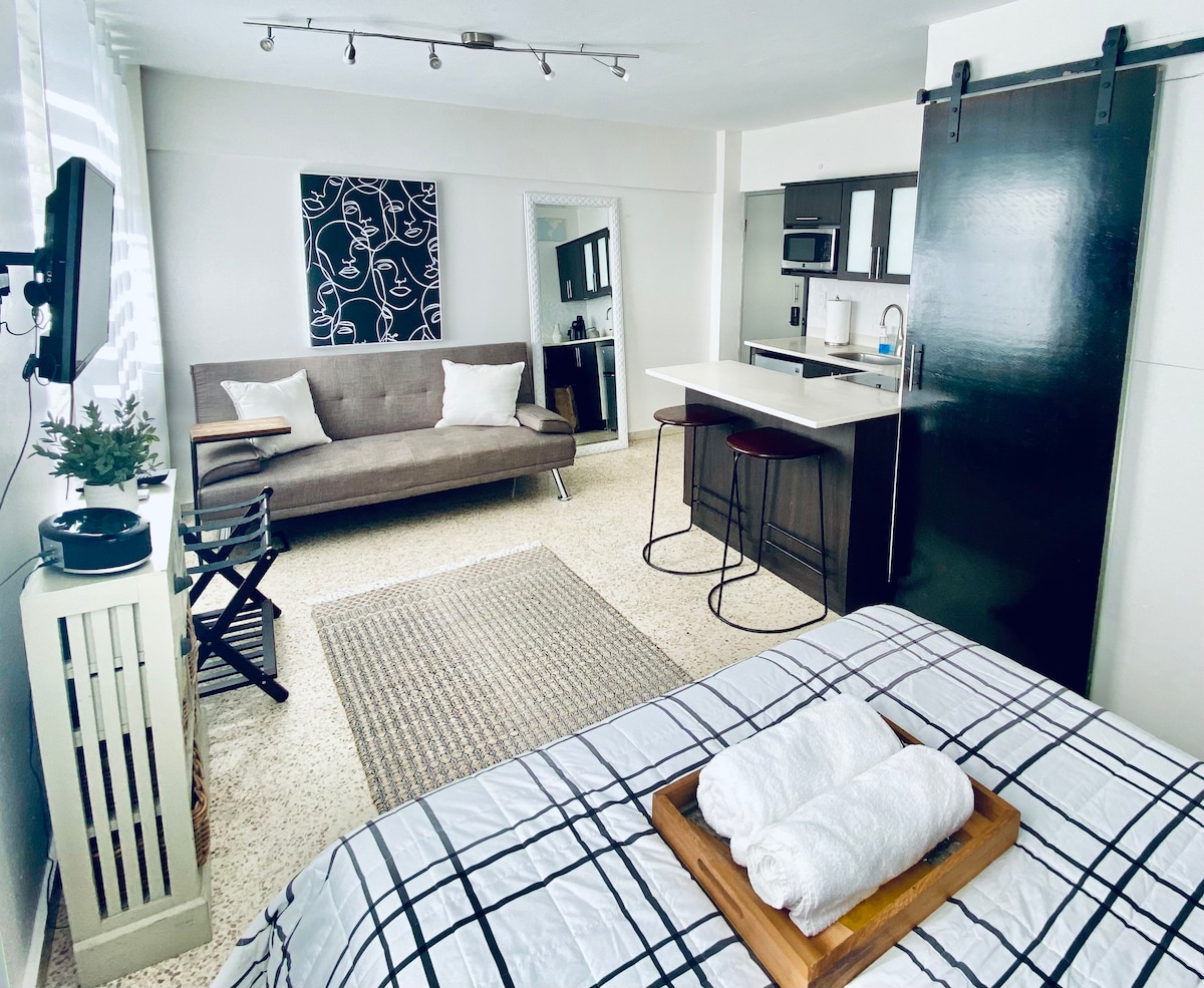 A cozy studio apartment features a spacious sleeping area with a checkered blanket and neatly arranged towels. A stylish kitchenette and dining space are visible, along with a modern sofa and a small table. Natural light illuminates the room through large windows.
