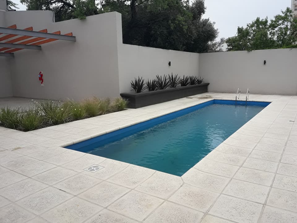 A private outdoor area is highlighted by a rectangular swimming pool, featuring blue water and steps leading in. Surrounding the pool are neutral-colored tiles and landscaped sections with greenery, creating a refreshing space for relaxation.