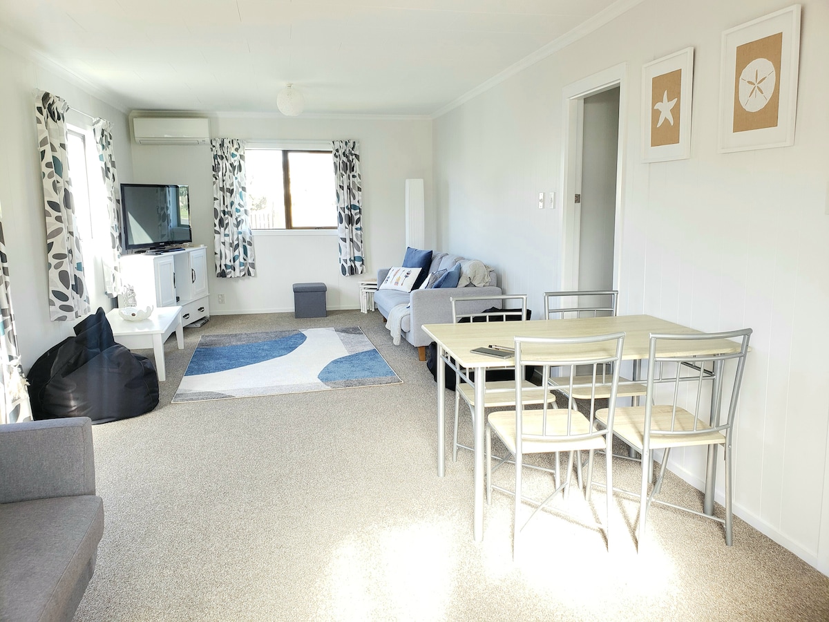 A spacious living area is featured, showcasing a grey sofa, bean bags, and a television. Natural light filters through large windows adorned with patterned curtains, while a dining table with six chairs occupies one corner. A soft area rug adds warmth to the neutral-toned room.