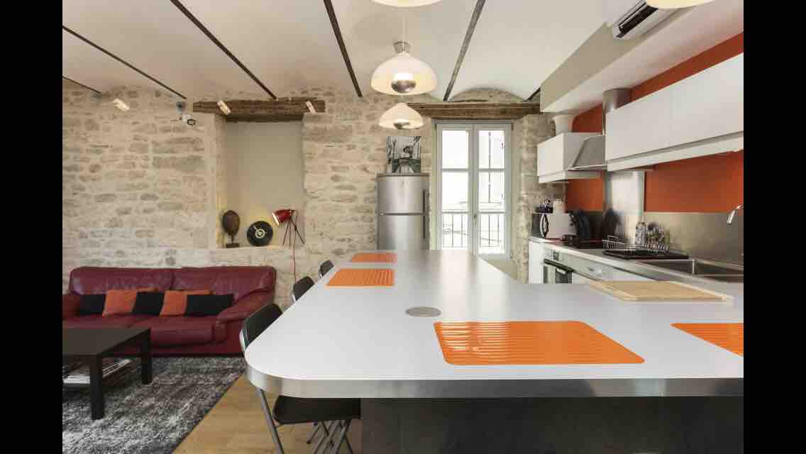A modern kitchen area is showcased, featuring a sleek white countertop with orange accents and black chairs. A spacious living area is visible, with a red sofa and a textured rug. Natural light floods in through large windows that enhance the stone wall ambiance.