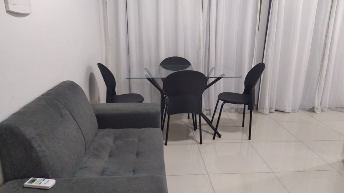 A modern seating area is presented, featuring a gray sofa positioned beside a clear glass dining table. Four black chairs surround the table. Sheer white curtains allow natural light to soften the space, complementing the polished tile floor.