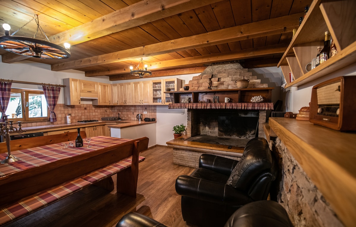 A spacious kitchen and dining area feature wooden cabinetry and a large dining table with a checkered tablecloth. A traditional stone fireplace is positioned against one wall, with comfortable leather seating nearby. Natural light enters through the window, complemented by rustic decor elements.