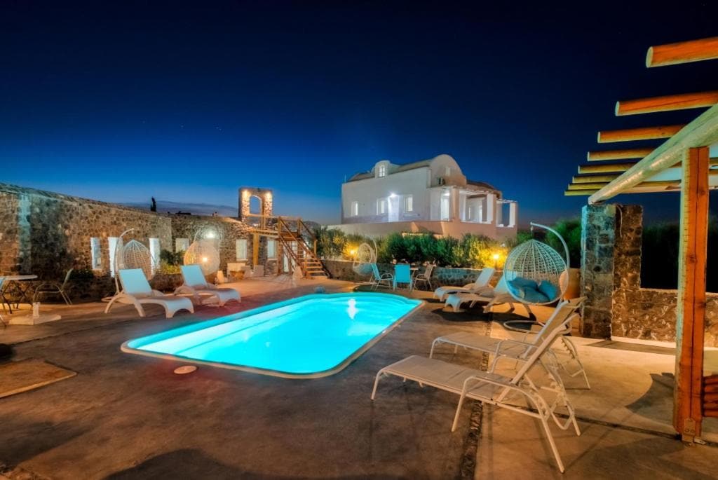 A spacious outdoor area is depicted, featuring a rectangular swimming pool surrounded by lounge chairs. Soft lighting enhances the atmosphere, and a two-story building is visible in the background. Hammock-style seating adds a relaxed vibe, complemented by stone walls and greenery.