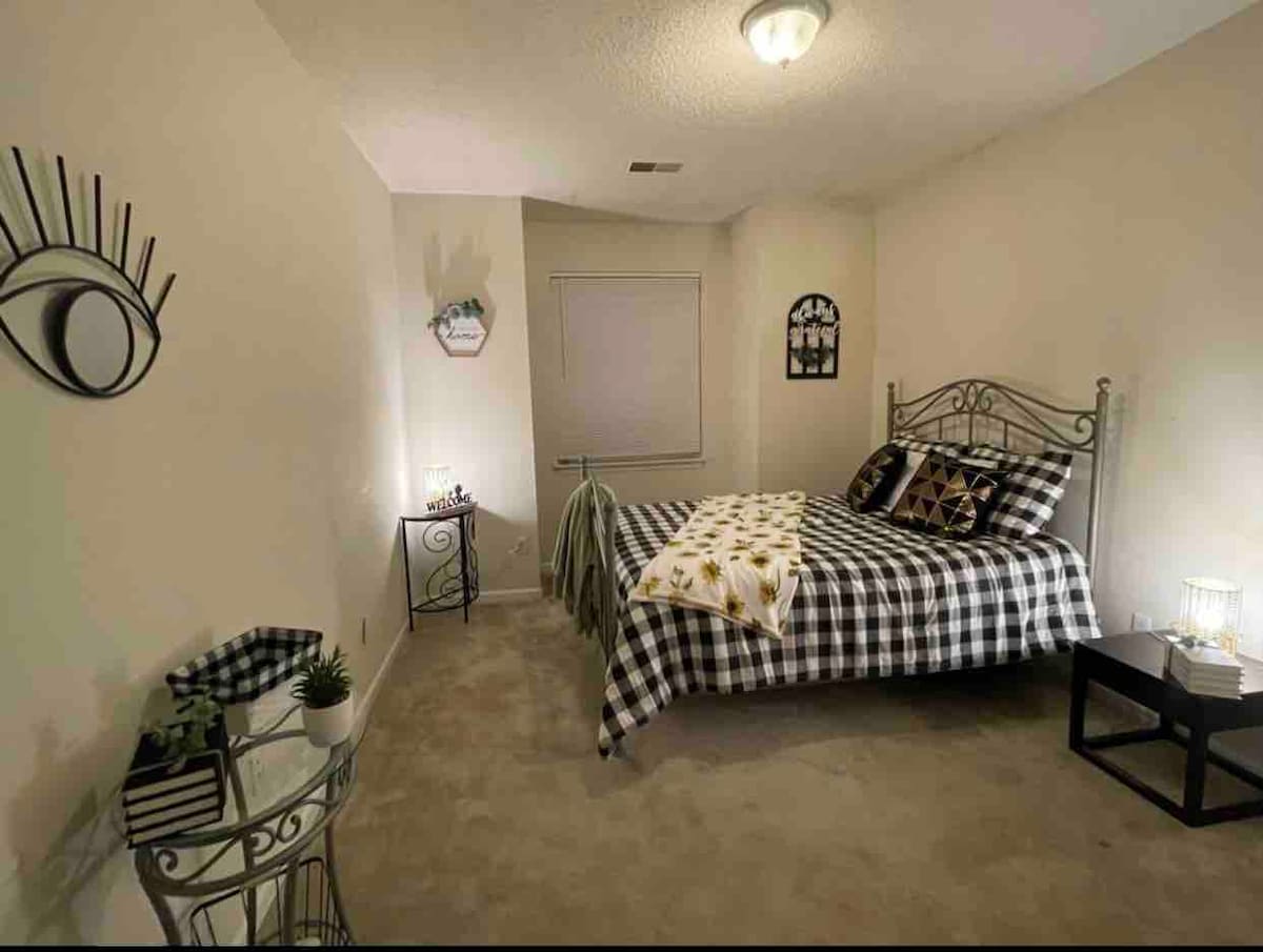 A softly lit bedroom features a spacious bed with a checked blanket and decorative pillows. A small table with a lamp sits beside the bed, and a window offers natural light. Decorative wall art and a plant add a touch of character to the room.