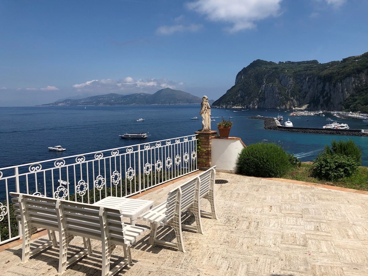A scenic terrace offers views of the coastline and the sea, with boats gently floating in the water. White lounge chairs are arranged around a stone table, creating a relaxed outdoor space. A statue stands elegantly, complementing the picturesque surroundings.