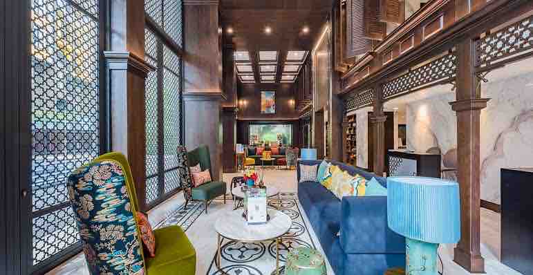 A spacious lounge area features high ceilings and intricate woodwork. Comfortable seating options include patterned armchairs and a blue sofa, arranged around a circular coffee table. Large windows provide ample natural light, while decorative elements and plants enhance the space.