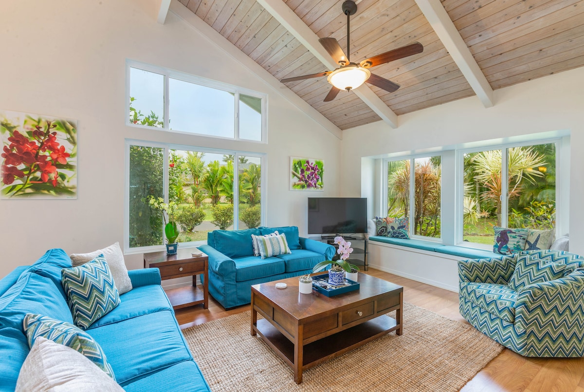 The spacious living area features high ceilings and large windows that allow natural light to illuminate the newly upholstered furniture. A television is positioned on a wooden media console, while tropical-themed artwork adorns the walls, adding to the serene ambiance.