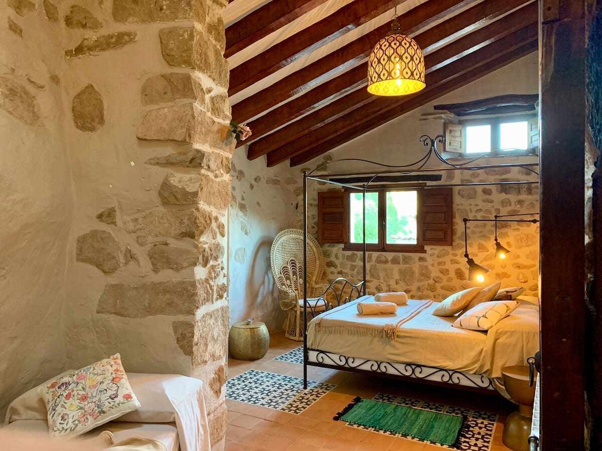 A warm and rustic room features a four-poster bed with neutral linens and decorative pillows. Exposed wooden beams enhance the charm, while a cozy seating area is present. Natural light enters through a window adorned with wooden shutters, complementing the stone walls.