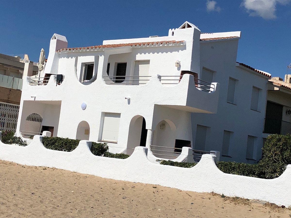 A white, two-story building is seen, featuring multiple balconies and windows. The property is prominently positioned near the beach, enclosed by a low white wall. The surrounding area indicates a tranquil environment, complemented by a sandy beach visible in the foreground.