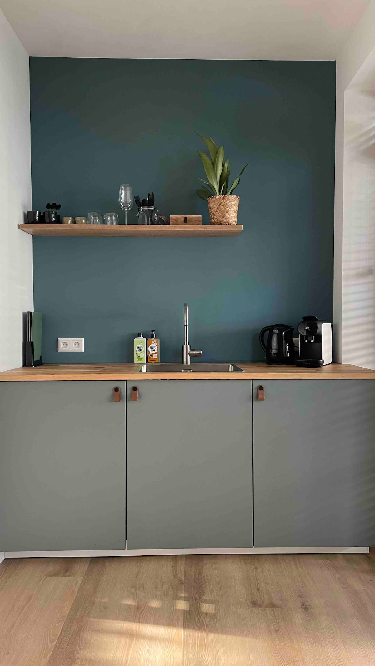 A compact kitchenette features a grey cabinet beneath a wooden countertop. A dark green wall serves as a backdrop, adorned with a small shelf holding glasses and kitchen items. A coffee machine and other appliances are positioned on the countertop, complemented by a potted plant.