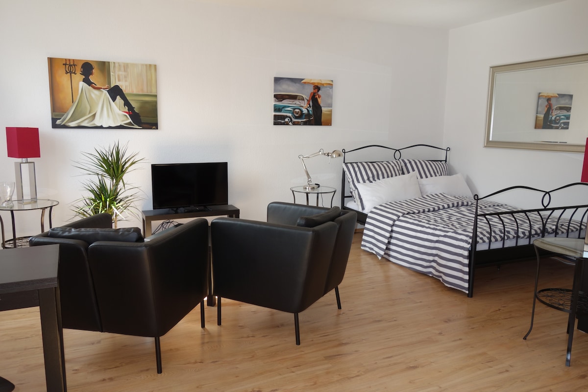 A spacious living area is visible, featuring a striped bed and two black armchairs. A contemporary television is situated on a stand. Decorative plants and framed artwork adorn the walls, complemented by a mirror reflecting the light color scheme and wood flooring.