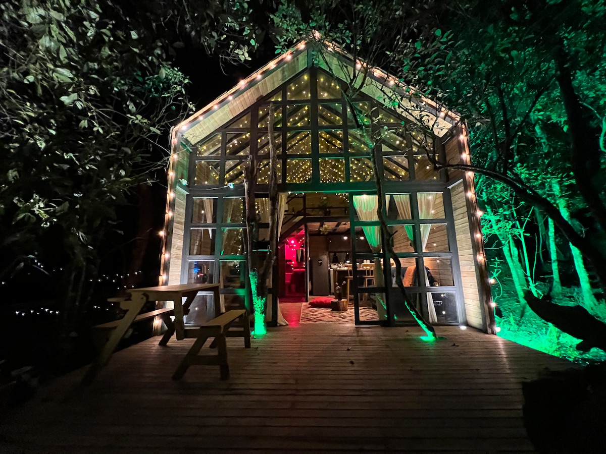 A charming glass-fronted chalet is illuminated by warm lights, creating a cozy ambiance. Wooden decking leads to an inviting dining area with a picnic table. Lush greenery encircles the structure, enhancing the natural setting. Inside, the space reveals a welcoming atmosphere with soft lighting.