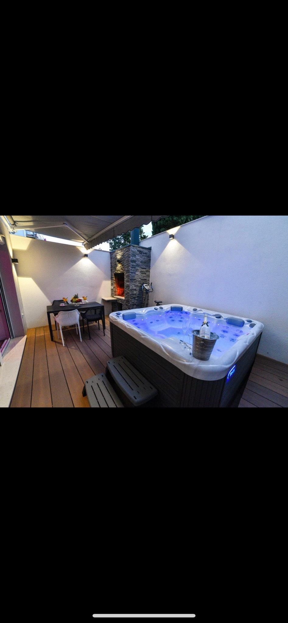 A private terrace is featured, showcasing a spacious hot tub with subtle blue lighting. A dining table is positioned nearby, accompanied by comfortable chairs. The setting is enhanced by ambient lighting along the walls, providing a serene atmosphere for relaxation.