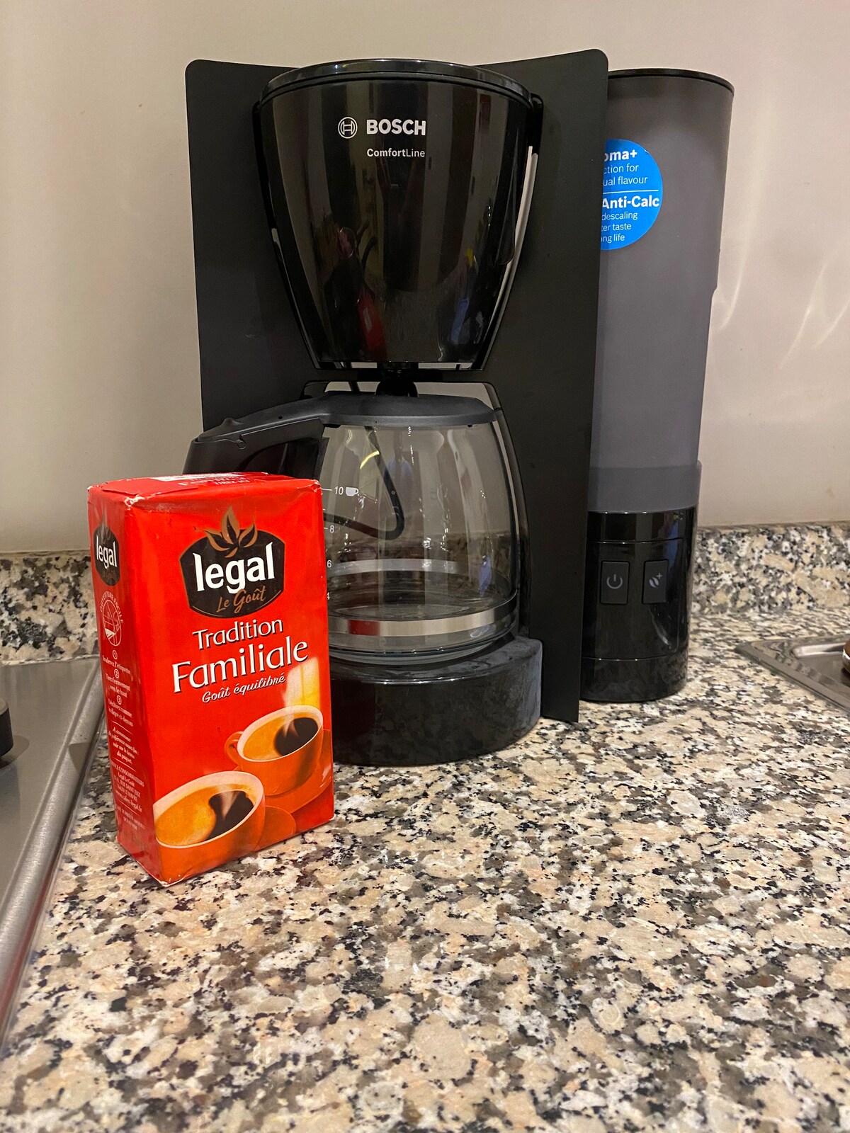 A German Bosch coffee machine is positioned on a granite countertop, accompanied by a box of coffee. The machine features a black and silver design, with a glass carafe to hold brewed coffee. Nearby, kitchen utensils are visible, indicating a fully equipped kitchen.