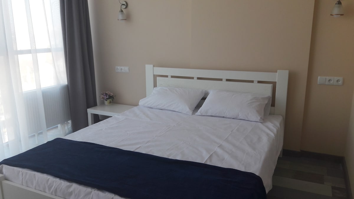 A bright and airy space features a spacious bed with white linens and two pillows. Natural light filters through sheer curtains, illuminating the room. A small table is placed beside the bed, with a delicate vase of flowers adding a subtle touch.