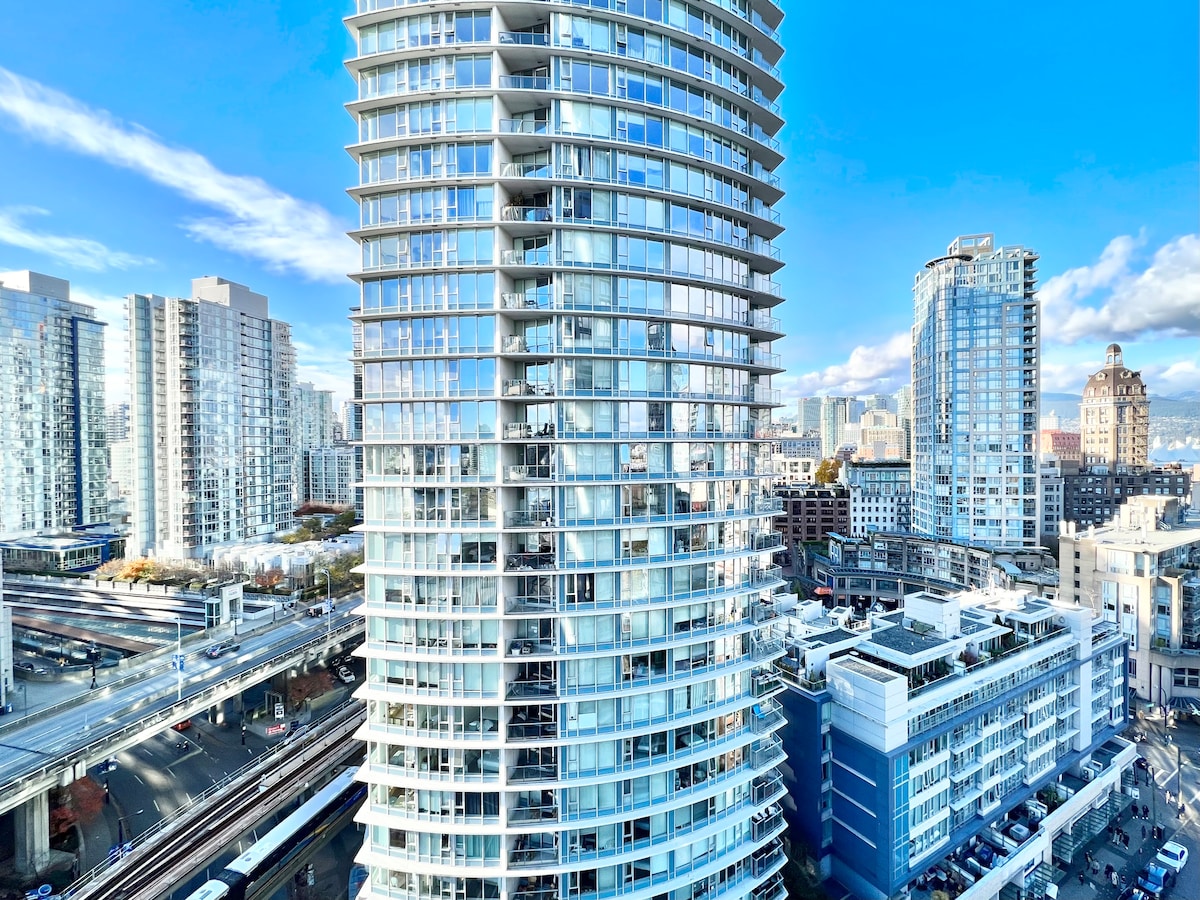 A modern, multi-story building with a circular design features extensive glass windows reflecting the surrounding cityscape. Nearby structures display various architectural styles, contributing to the urban environment. The clear blue sky adds to the vibrant atmosphere of the scene.