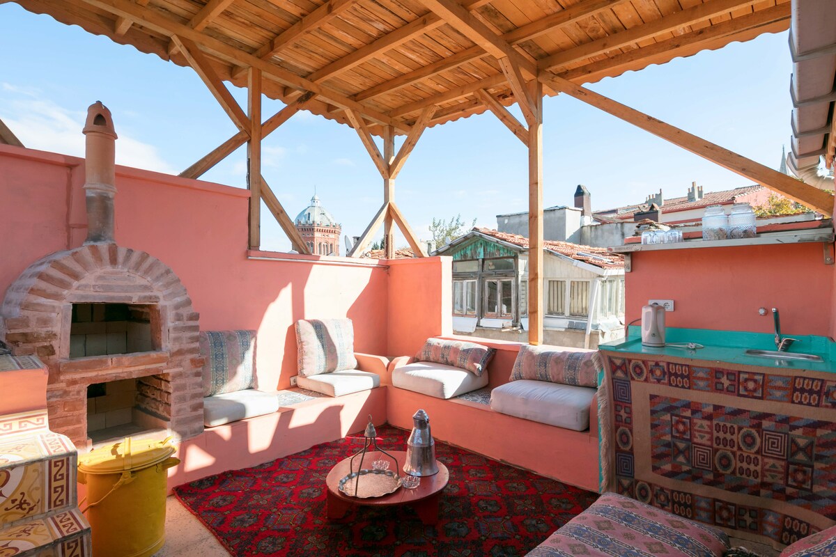 A communal terrace is showcased, featuring built-in seating with cushions around a low table. A traditional Turkish coffee pot and glasses are on the table. A brick oven is visible, and the area is sheltered with wooden beams, offering views of the neighboring rooftops.