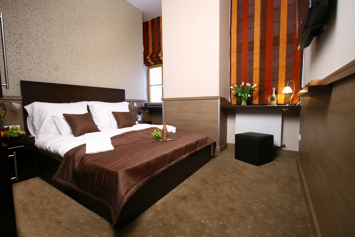 A well-appointed guest room features a spacious bed with dark brown bedding and white pillows. A small seating area is visible, alongside a desk with a lamp and a vase of flowers. Natural light enters through a window dressed with striped curtains.