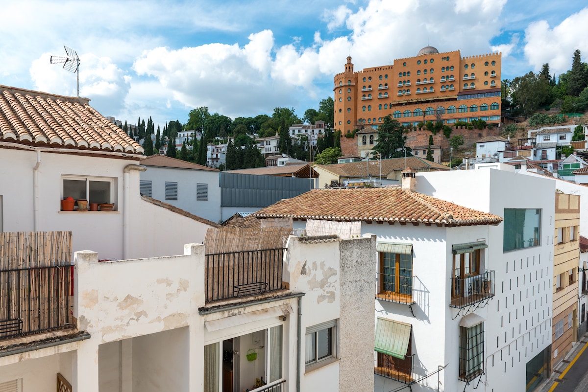 A view of neighborhood rooftops, showcasing a mix of traditional and modern architecture. A prominent hillside building is visible in the background, framed by a clear blue sky with scattered clouds. The scene conveys the community's blend of charm and urban life.