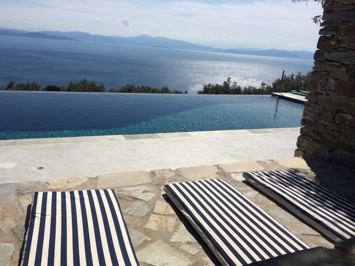 An infinity pool extends towards the horizon, reflecting the sunlit sea. Three striped lounge chairs are positioned on a stone terrace, providing a serene spot to relax. The calm water and distant hills create a tranquil backdrop.