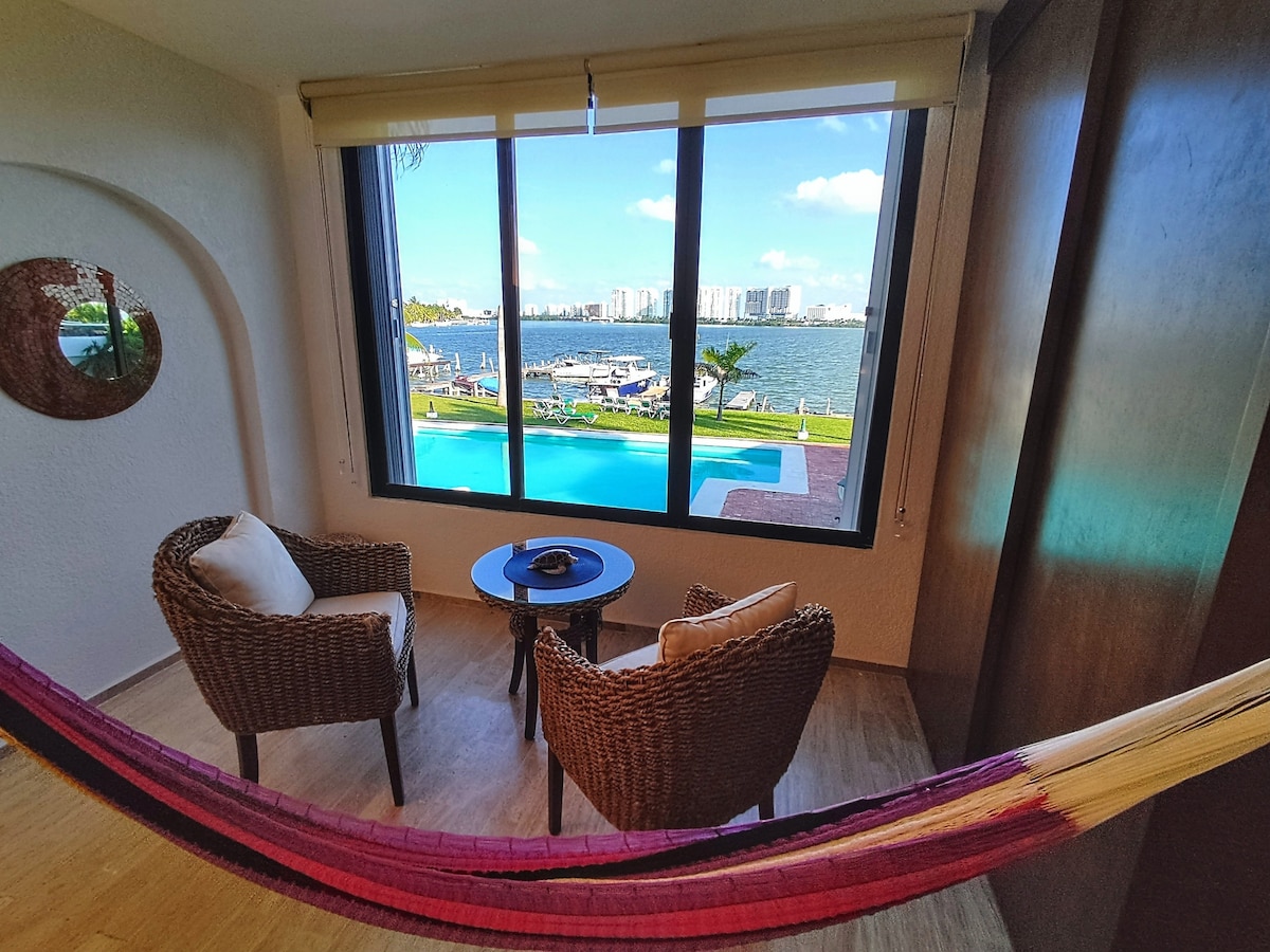 A serene seating area is positioned near large windows, offering views of a swimming pool and lagoon. Two woven chairs are accompanied by a small table. A colorful hammock adds a touch of comfort, enhancing the space's inviting atmosphere.