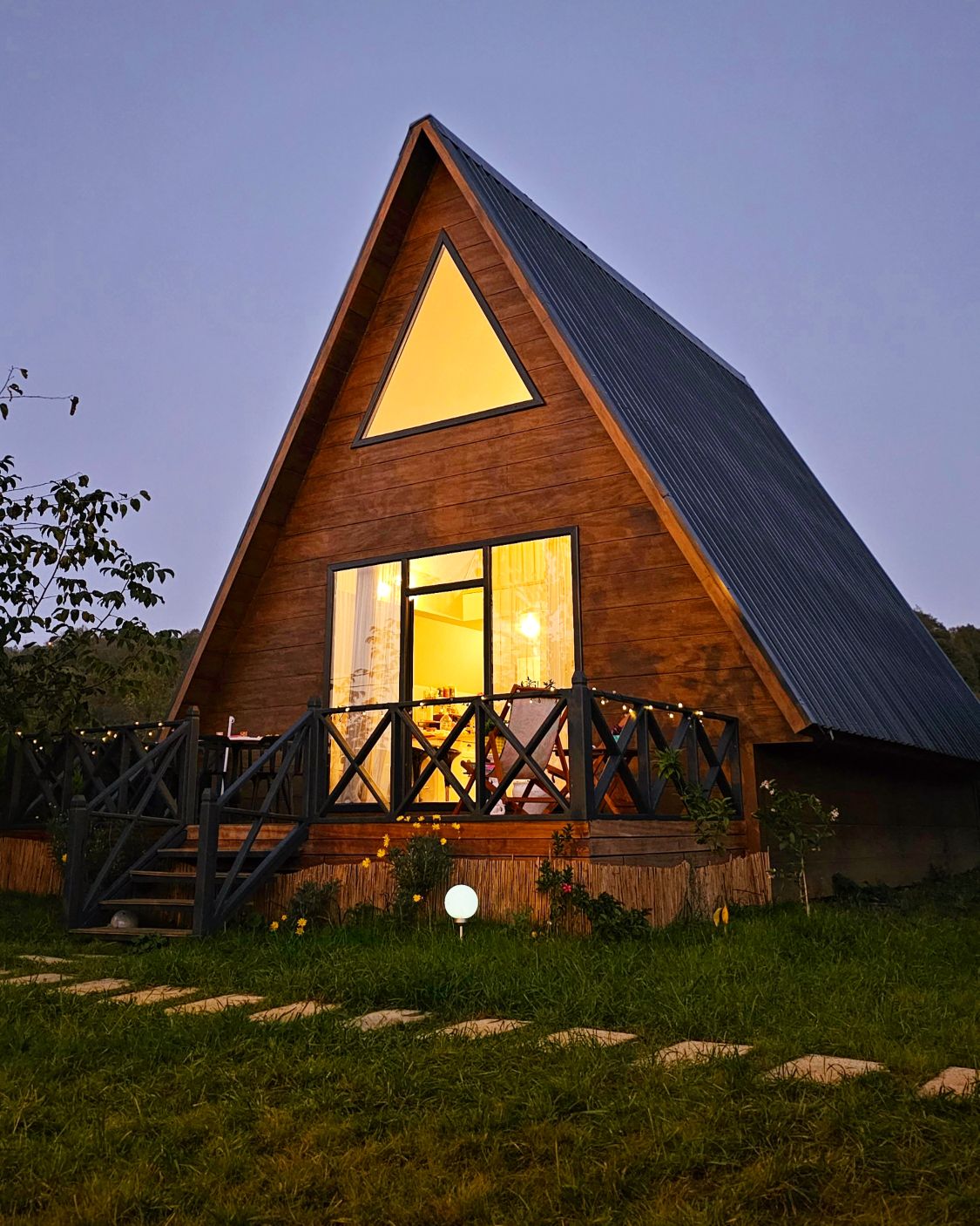 A triangular wooden cabin is set among greenery, featuring large windows that reflect warm lighting. A wraparound balcony with a railing provides outdoor space, while a pathway of stones leads to the entrance, inviting guests to enjoy the natural surroundings.