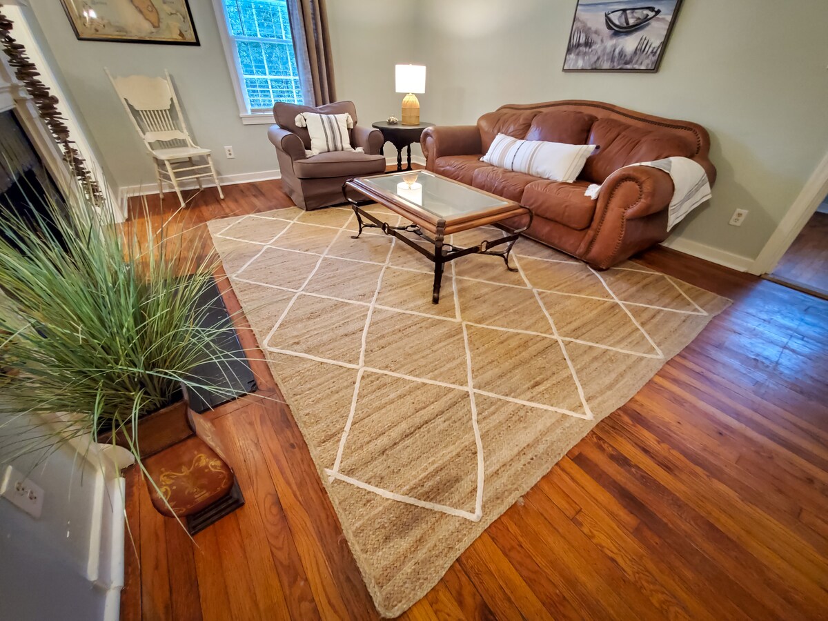 A cozy living area features a plush brown sofa and an accent chair, complemented by a glass-top coffee table on a geometric-patterned area rug. Natural light enters through nearby windows, creating a warm environment while wooden floors add a touch of elegance.