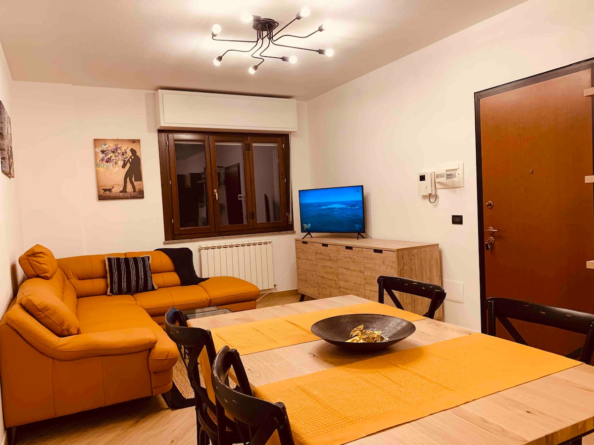 An open living area is featured, equipped with a modern orange sectional sofa and a wooden dining table. A flat-screen TV is positioned against the wall, and natural light enters through a window. Decorative elements include a central bowl on the table and art on the wall.