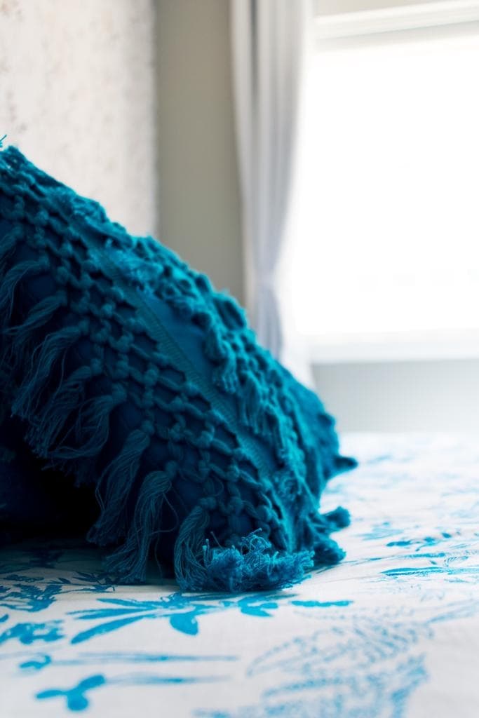A textured turquoise pillow is positioned on a bed with a white and blue patterned cover. Sunlight filters through a nearby window, creating a bright and airy atmosphere in the room.