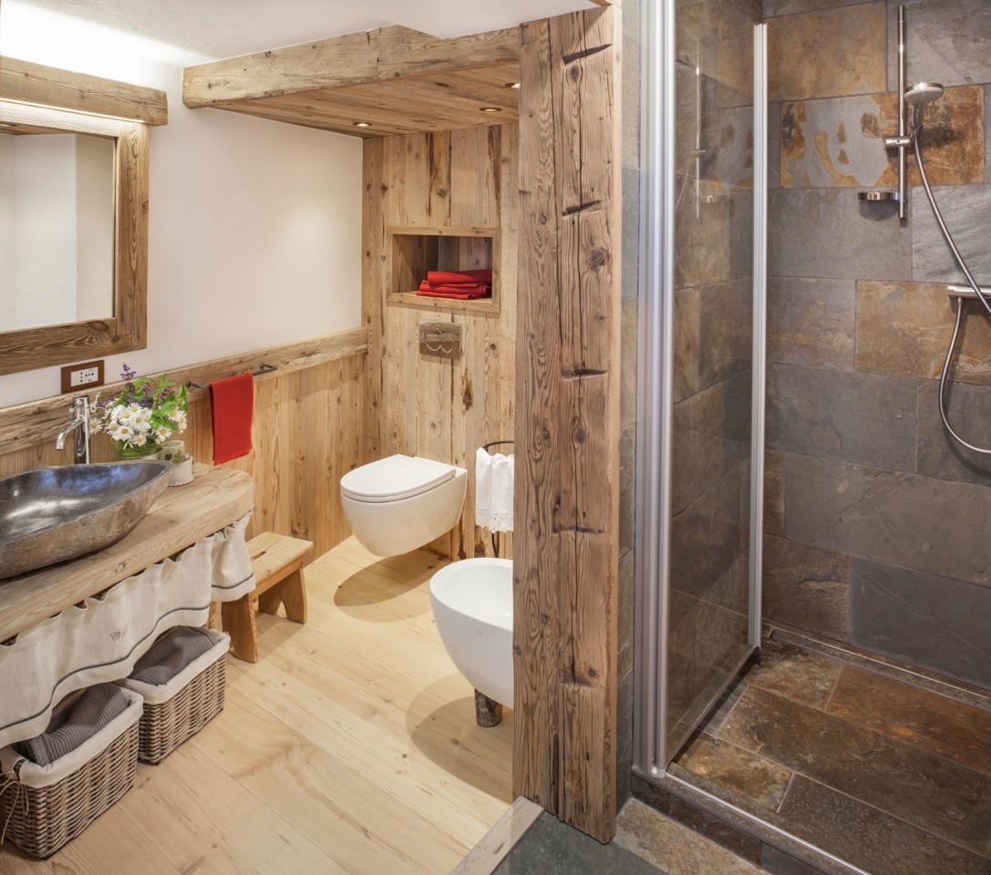A natural wood bathroom is showcased, featuring a wooden sink with a stone basin. A shower with a glass door stands nearby, with slate wall tiles adding a rustic touch. Soft linens are neatly arranged, complemented by red towels on a shelf.