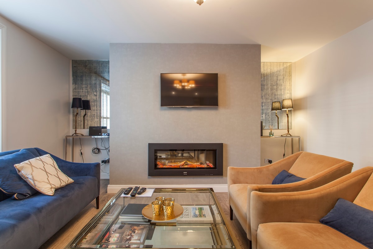 An open living area features a modern electric fireplace set within a wall. A large flat-screen TV is mounted above it. A glass coffee table is centrally placed, with two armchairs and a sofa providing comfortable seating, complemented by warm lighting from nearby lamps.