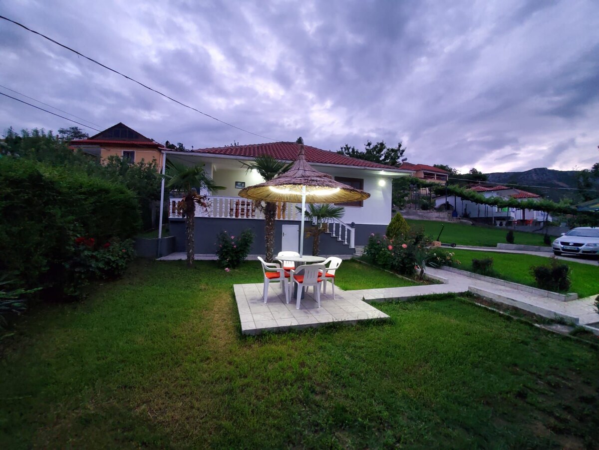 An outdoor dining area is positioned on a stone patio, featuring a table with four chairs and a large umbrella providing shade. Lush green grass surrounds the space, with shrubs and palm plants adding to the natural landscape. A peaceful home is visible in the background under an overcast sky.