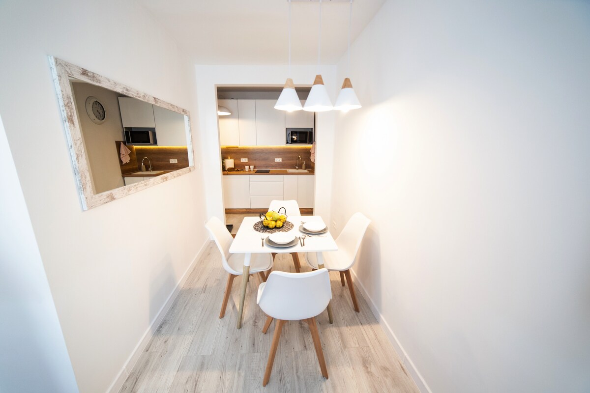 A bright dining area is featured, showcasing a white table surrounded by four sleek chairs. Soft lighting is provided by pendant lights above the table, and a mirror enhances the sense of space. A small bowl of fruit adds a touch of color.