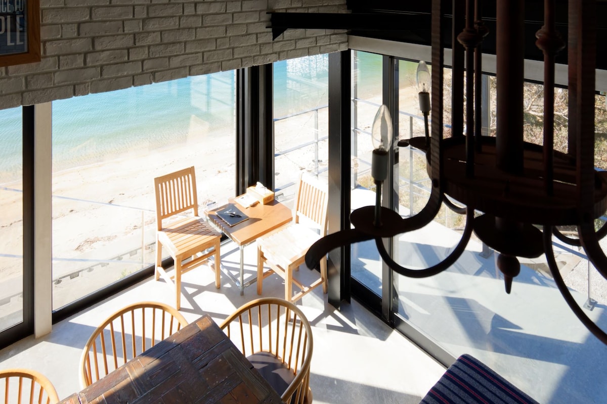A bright dining area is showcased with wooden chairs and a table, surrounded by large glass windows offering views of the sandy beach and serene water. Natural light enhances the inviting space, while suspended light fixtures complement the design.