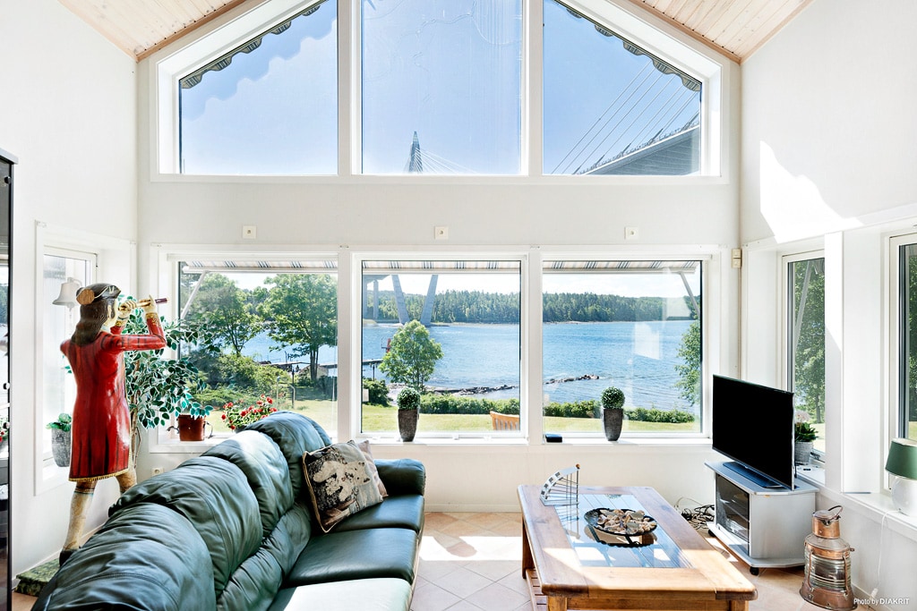 A spacious living area is highlighted by large windows that provide views of the waterfront. Sunlight fills the room, illuminating the seating arrangement and a coffee table. A decorative figure stands near a potted plant, enhancing the space's character.