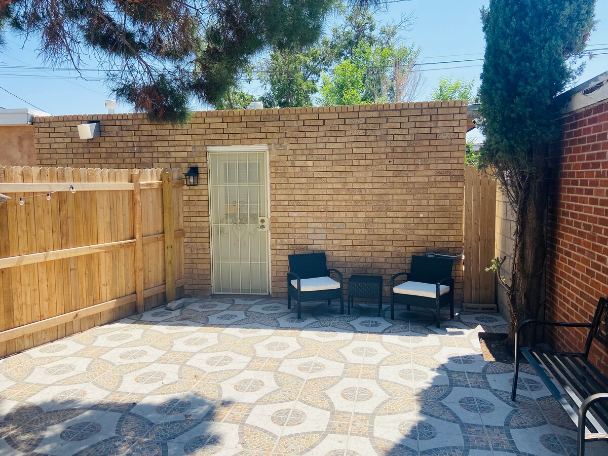 A private patio area is visible, featuring a patterned stone floor and surrounded by a wooden fence for privacy. Two chairs and a small table are positioned for seating. A door leads into the interior space, offering a cozy outdoor atmosphere.